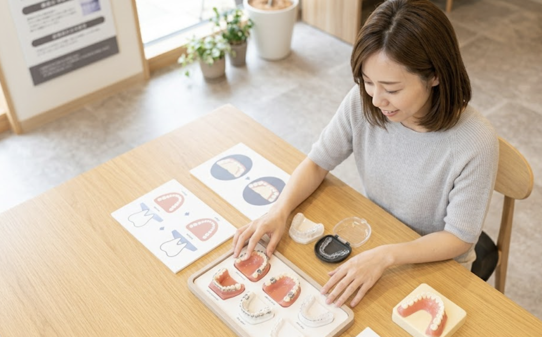 歯科医院の明るいプレゼンテーションエリアで、上から複数の治療方法の資料を覗き込む日本人女性の様子