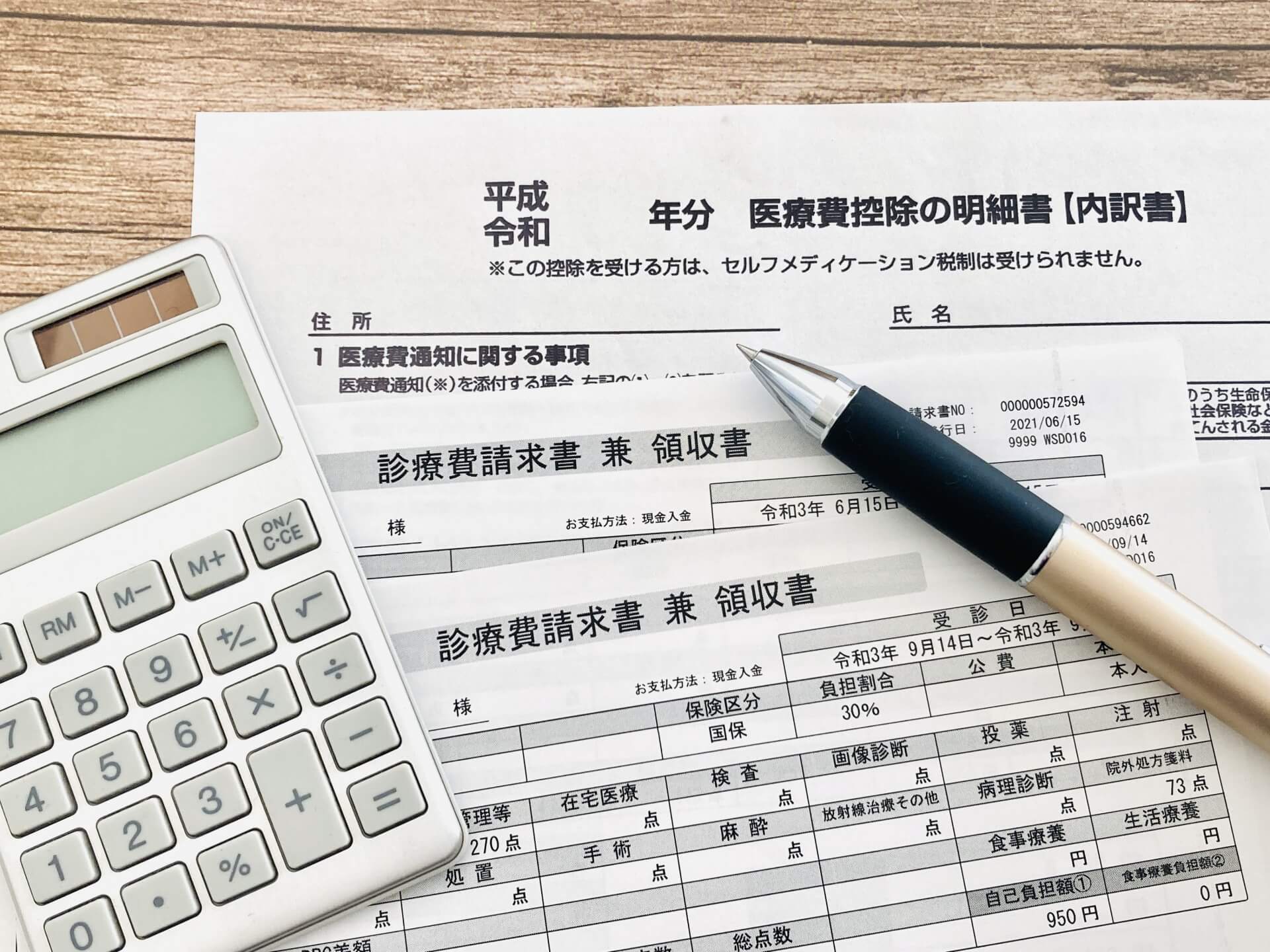 木目の机に置かれた医療費控除の明細書と診療費領収書、白い電卓、ボールペンの写真