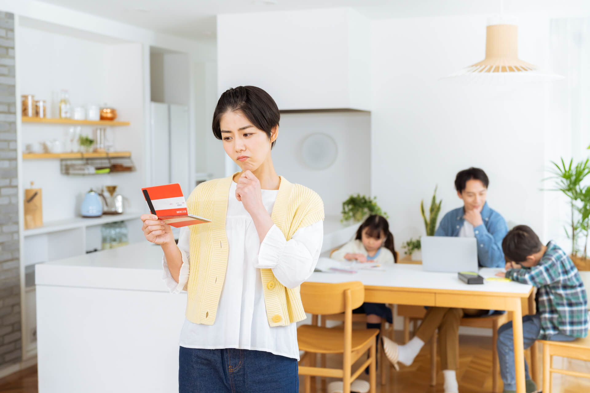 通帳を見て考え込む短髪の日本人女性。背景にダイニングの家族。